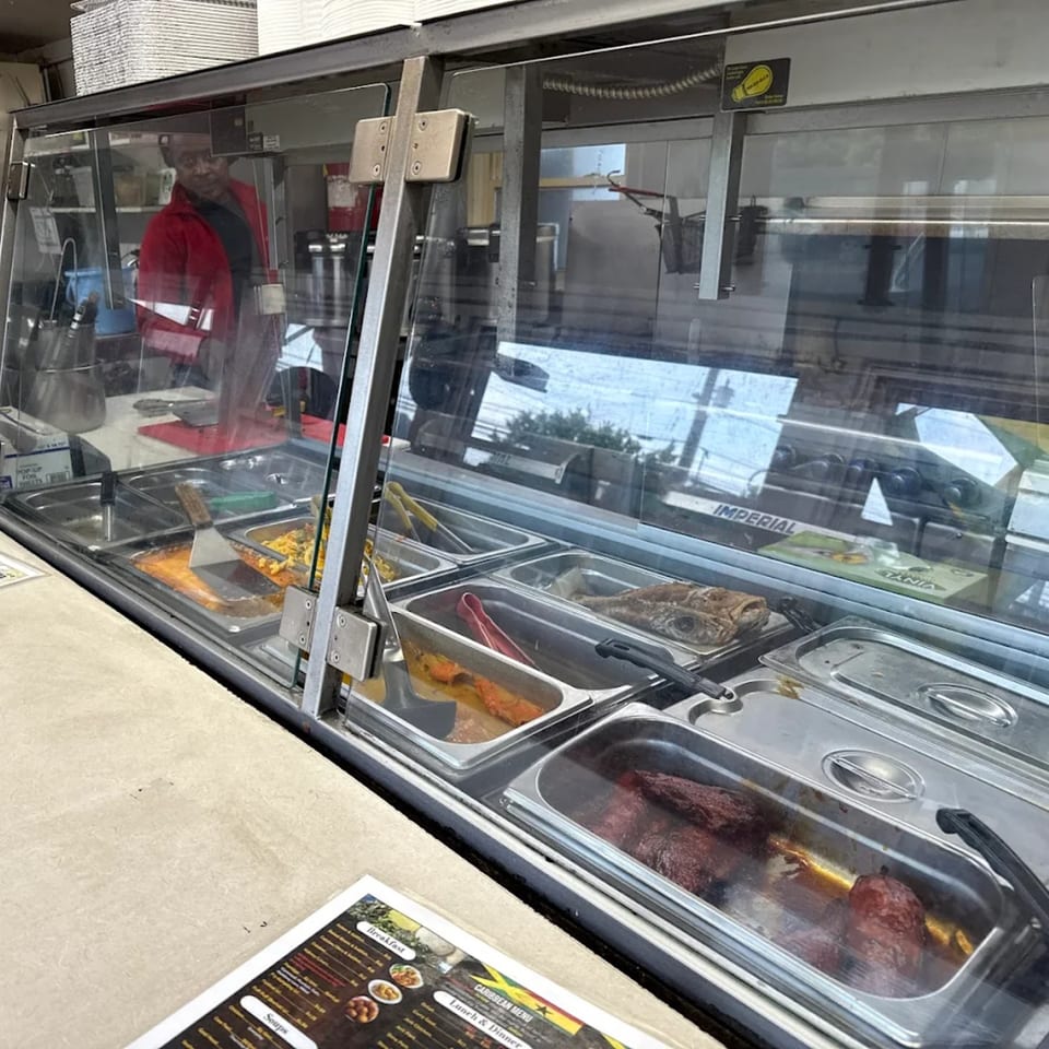 A server in a red jacket behind a hot food bar with various Caribbean dishes at OSU Jerk Grill.