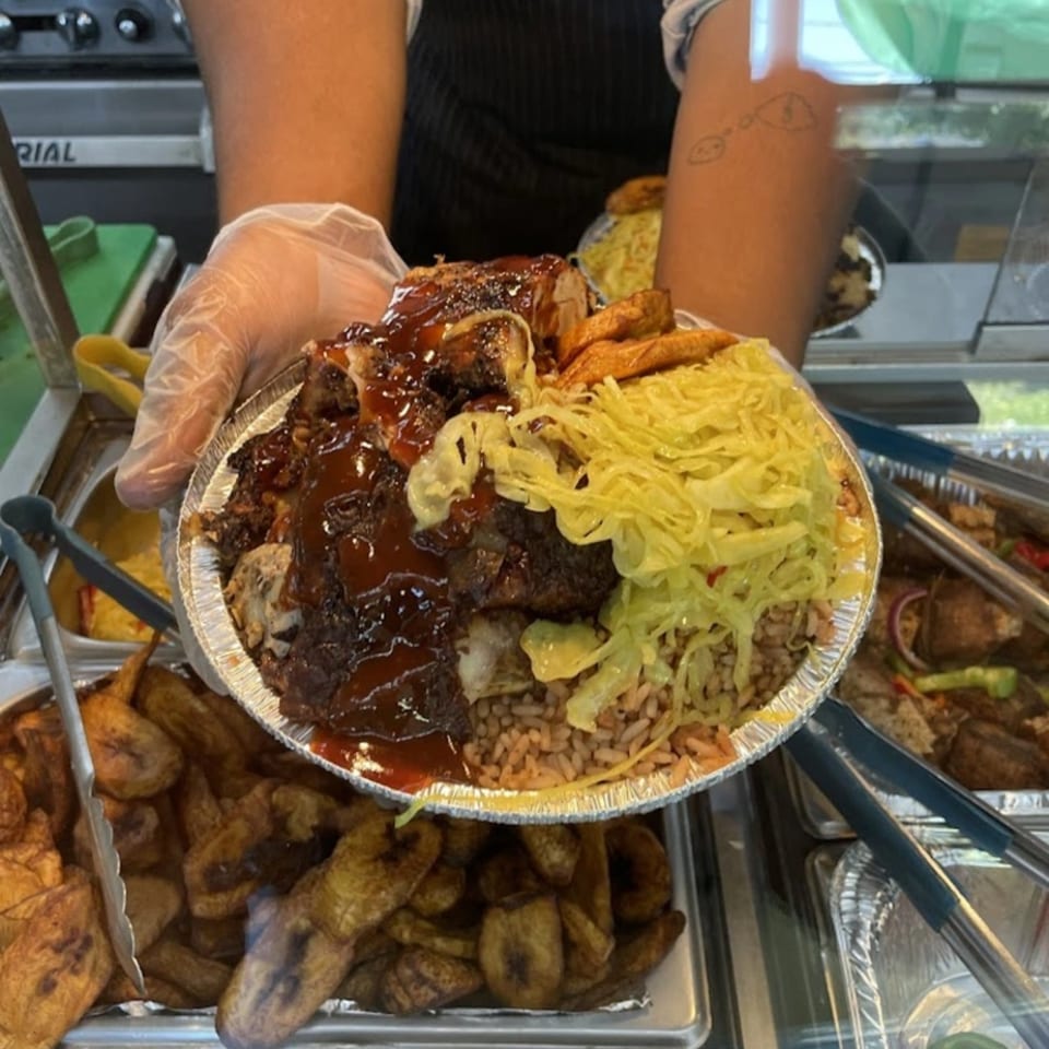 Gloved hands hold a foil plate of OSU Jerk Grill's jerk chicken, rice, cabbage, and plantains.