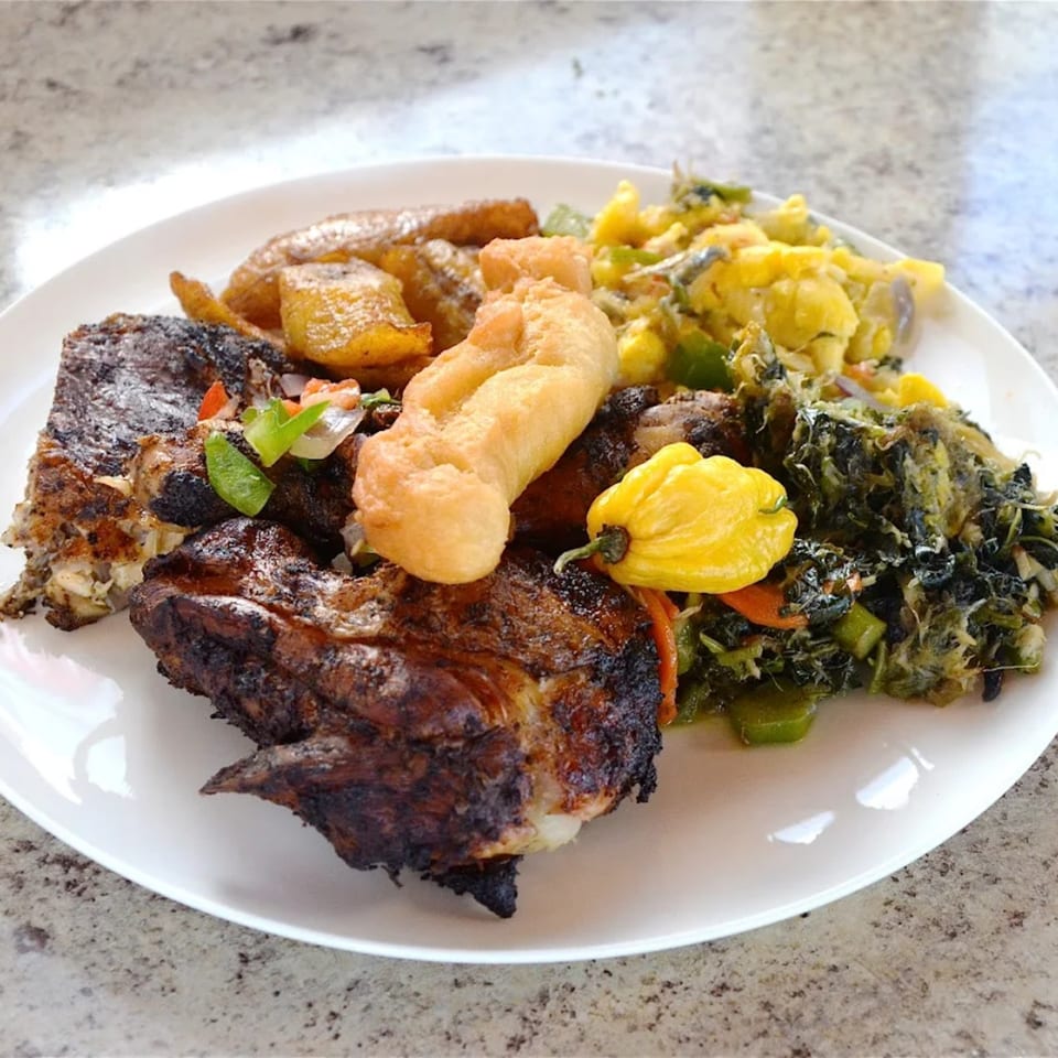 A white plate holds OSU Jerk Grill's jerk chicken, fried dumplings, plantains, and callaloo.