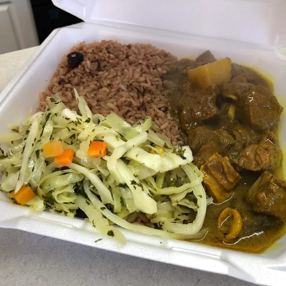 Curried goat with rice and peas, and steamed cabbage in a white takeout container.