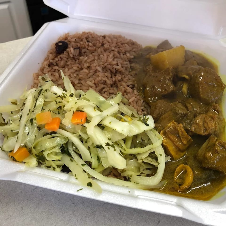 A Caribbean meal with brown rice, curried meat, and sautéed cabbage with carrots in a takeout container.