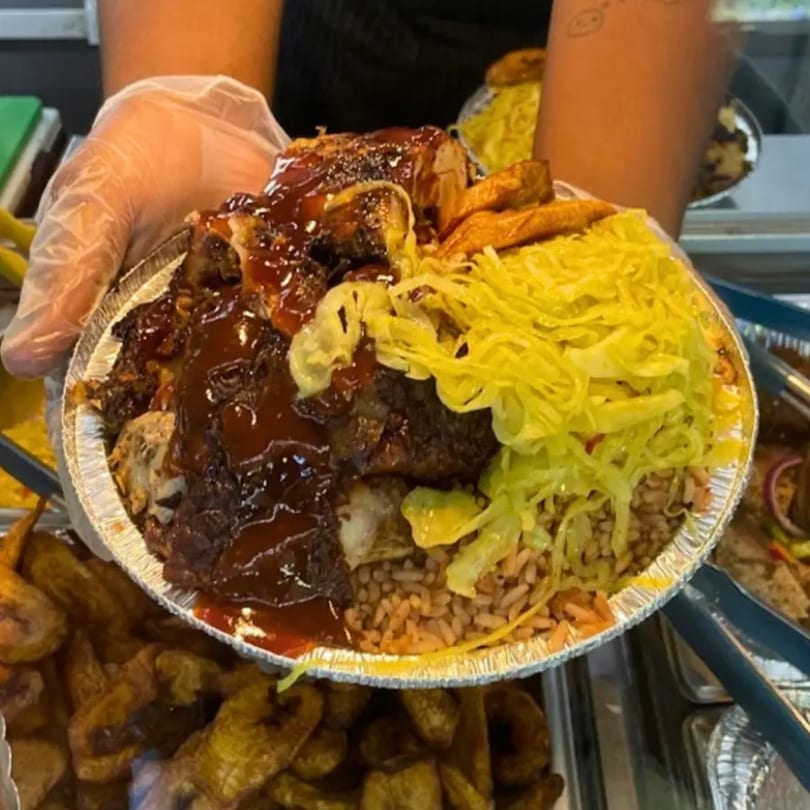 Gloved hands holding a foil container of OSU Jerk Grill's jerk chicken, rice, plantains, and cabbage.