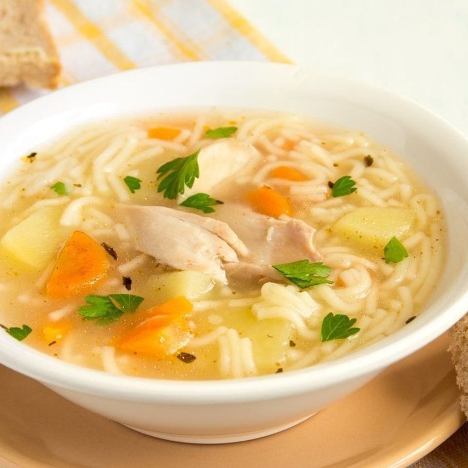 A white bowl of chicken noodle soup with carrots, potatoes, and parsley on a brown plate.