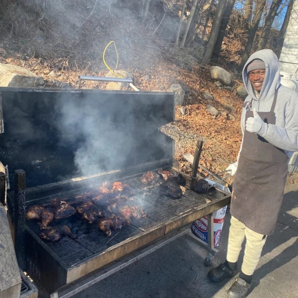 A man in a grey hoodie and apron gives a thumbs-up next to a large, smoking jerk grill.