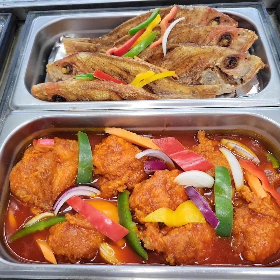 Two buffet trays of Caribbean food: fried fish with peppers and chicken in red sauce with vegetables.